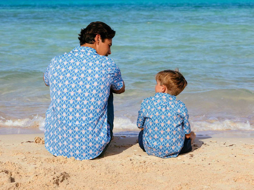 Father and son sitting on the beach wearing matching shirts.