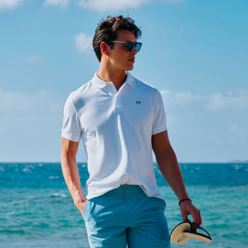 man standing in front of ocean in white skipjack polo and blue shorts